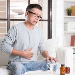 Mature 50s Asian man suffering heartburn or stomachache, pressing on stomach with painful expression, sitting on sofa at home, medicines on table