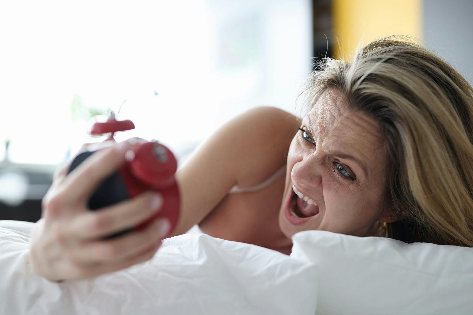 Woman lying in bed with alarm clock in her hands and screaming. Time management how to do everything concept
