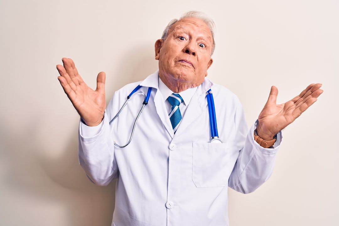 Senior grey-haired doctor man wearing coat and stethoscope standing over white background clueless and confused with open arms, no idea and doubtful face.
