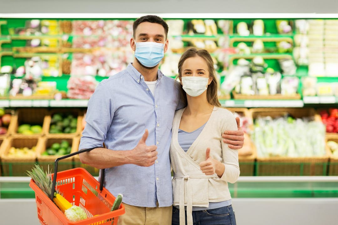 Shopping, sale, consumerism and people concept &#8211; happy couple wearing face protective medical masks for protection from virus disease with food basket at grocery store or supermarket showing thumbs up
