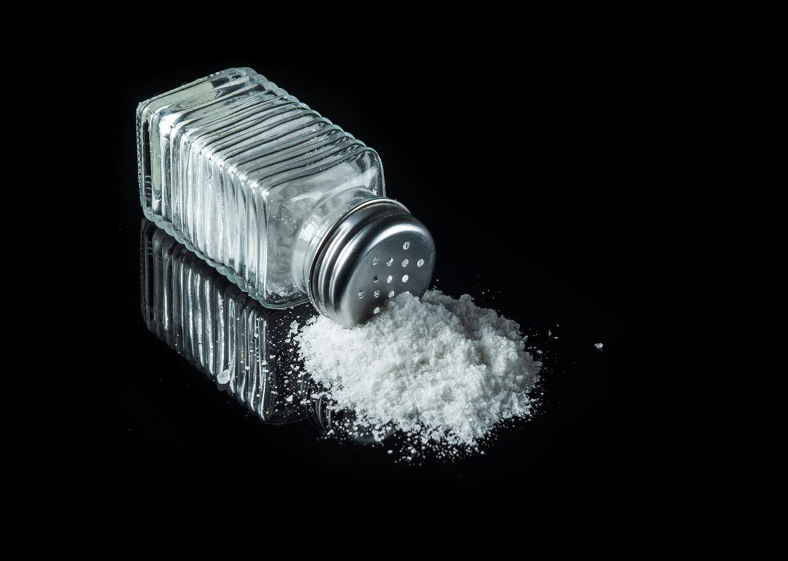 Heap of salt from a salt shaker on a black background. The concept of excessive salt intake that can be the cause of diabetes or other diseases
