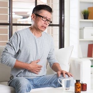 Mature 50s Asian man stomachache,  pressing on stomach with painful expression, sitting on sofa at home, medicines on table.
