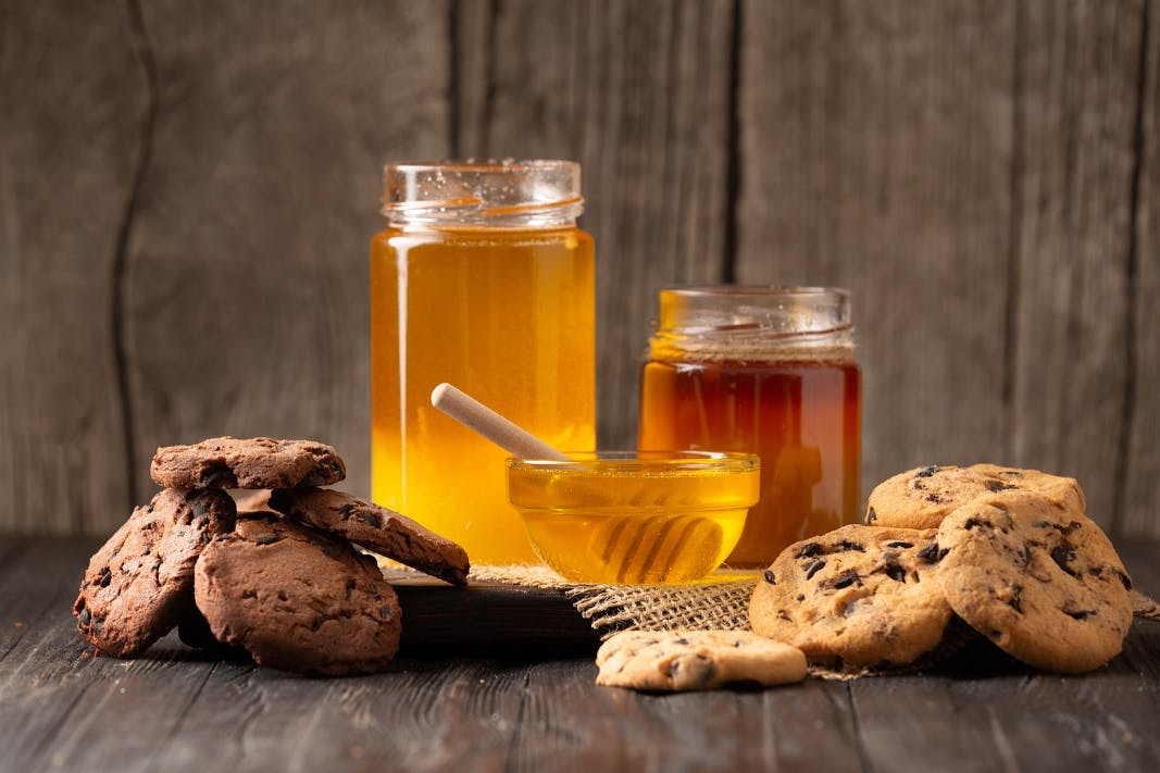 Honey in open jars and in a bowl, dipper and homemade cookies with chocolate chips on a wooden background. Homemade cakes and honey for breakfast. Composition of craft chocolate cookies and honey.
