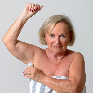 Close up Middle Aged Blond Lady Applying Deodorant on Armpit After Shower While Looking at the Camera Against Light Gray Wall.
