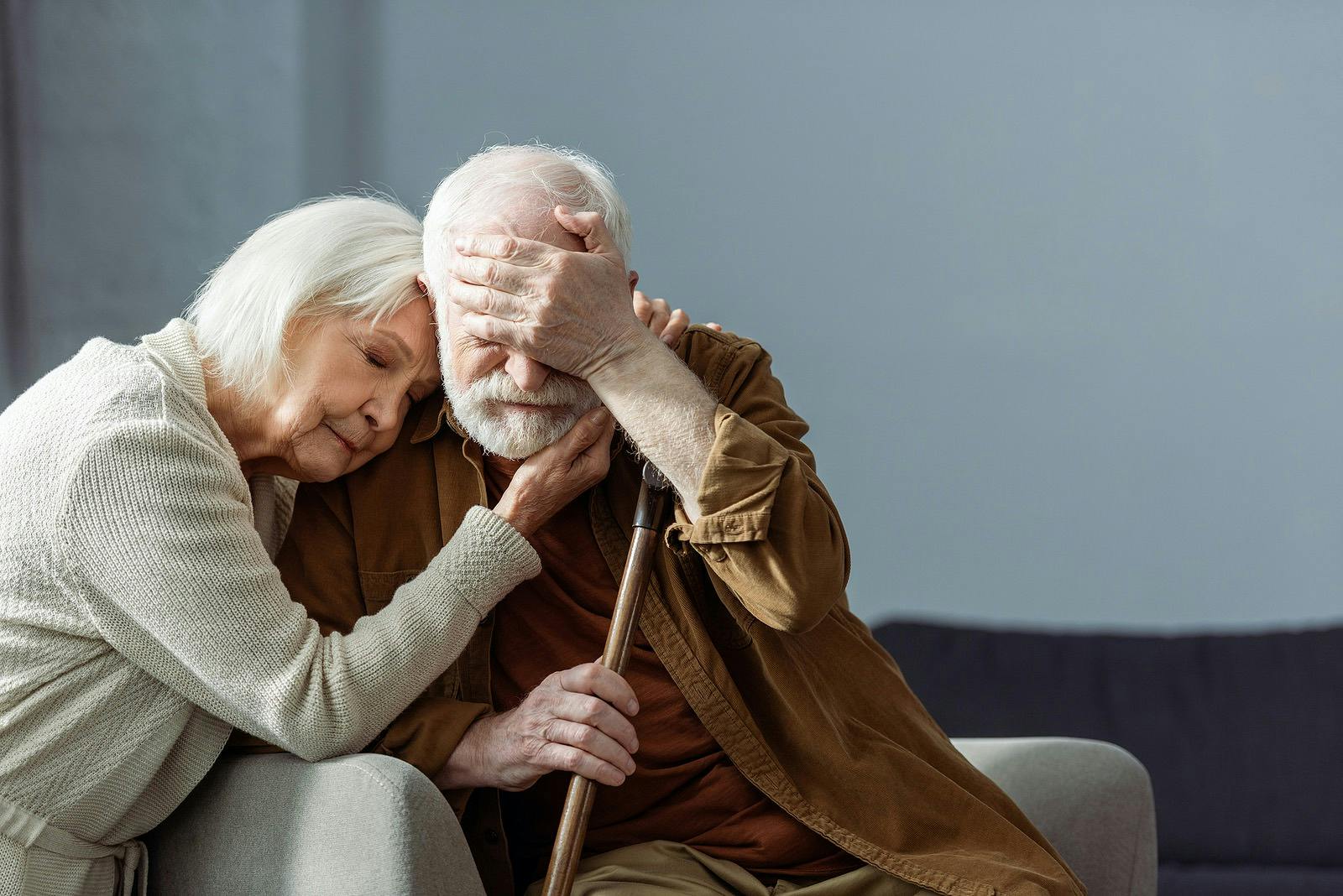 Senior man, sick on dementia, covering eyes with hand while wife embracing him with closed eyes
