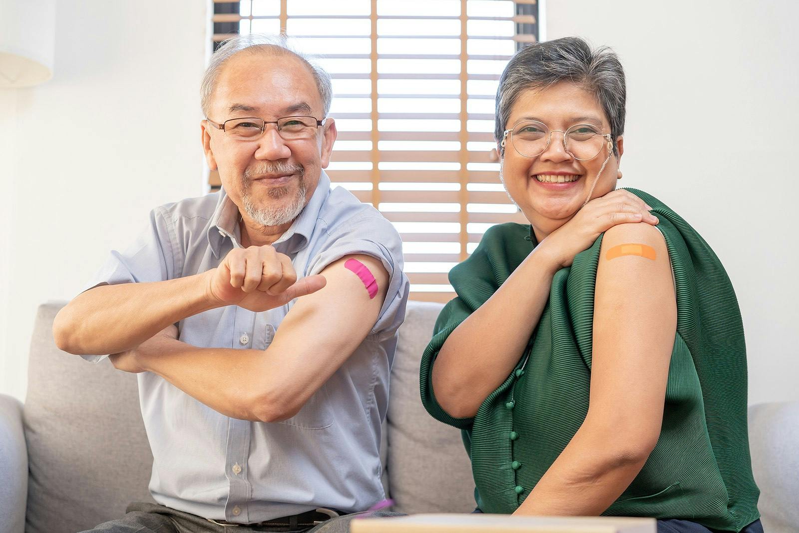 Coronavirus Vaccination, happy asian elderly, aged family smile strong together, showing bandage on arm with protect of covid-19 after injection of vaccine, sitting on couch in living room at home.
