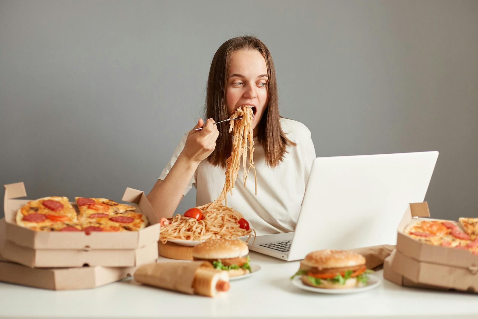 Indoor shot of busy hungry woman eating fast food while working on laptop, sitting at table against gray wall, working on laptop and eating pasta, keeps mouth open, looking at notebook display.
