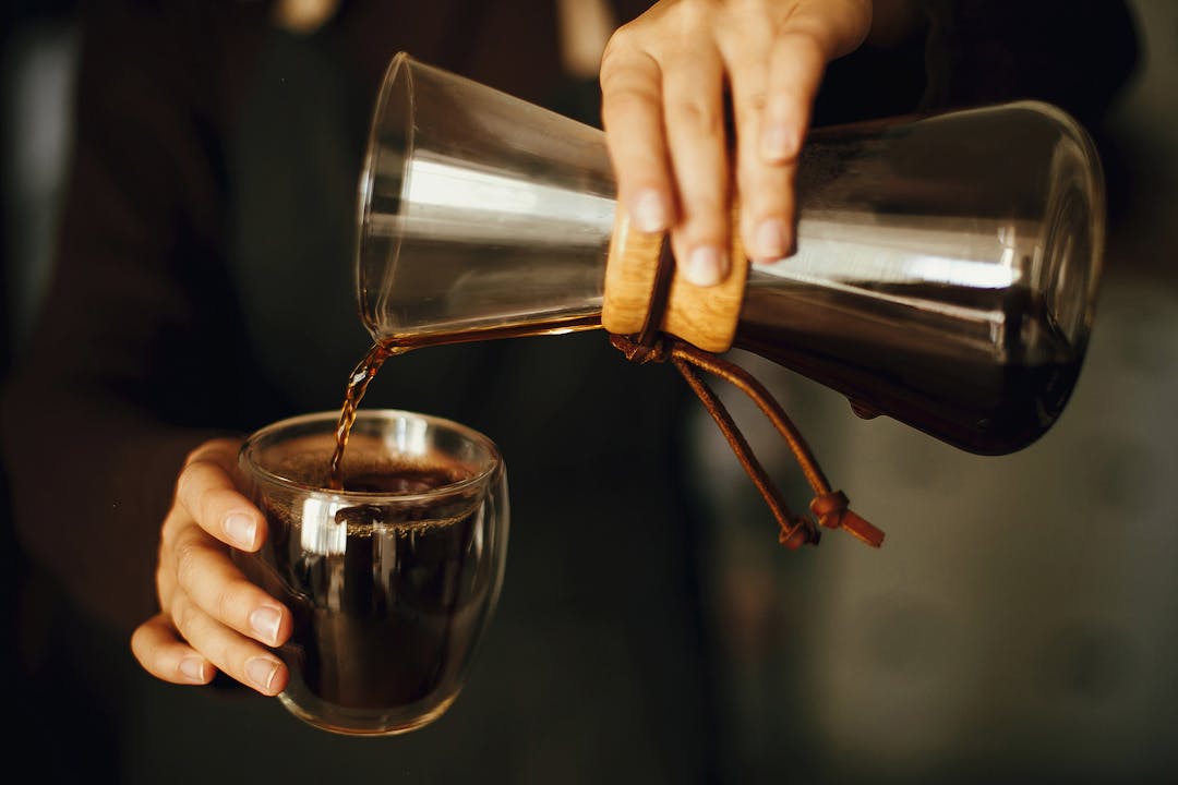 Professional female barista in black uniform making drip coffee. Person pouring fresh aromatic coffee from glass kettle in cup.  Alternative coffee brewing,
