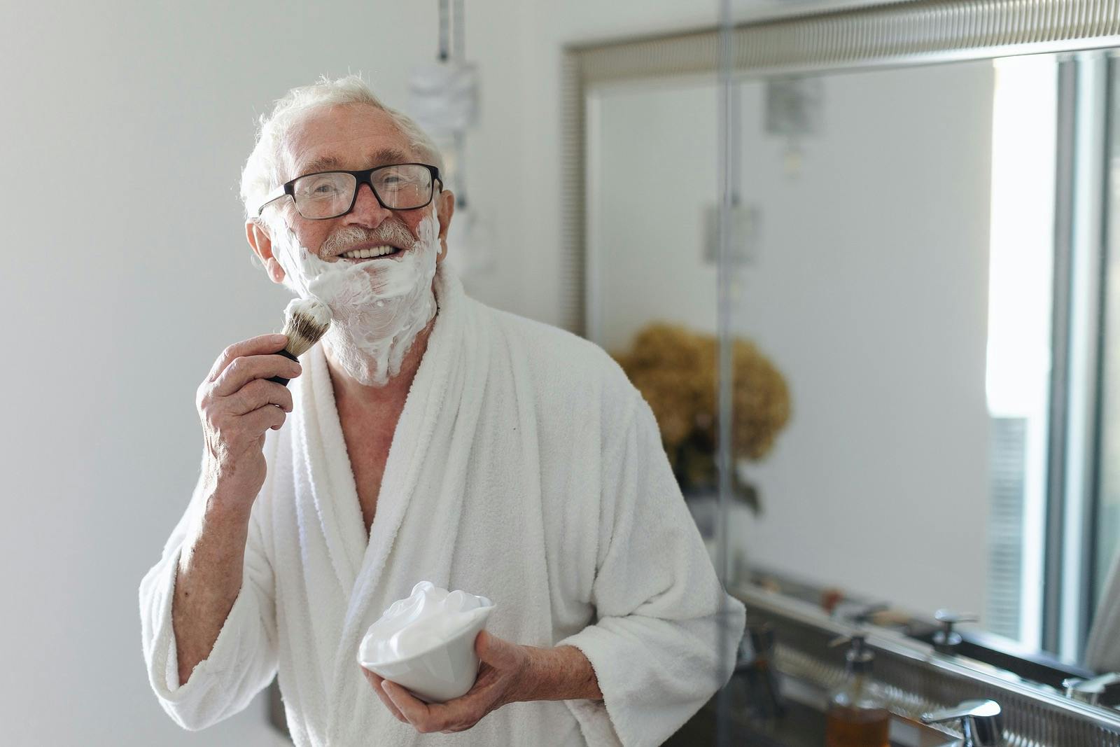 Senior man shaving his beard in the bathroom, looking at camera. Morning routine concept.
