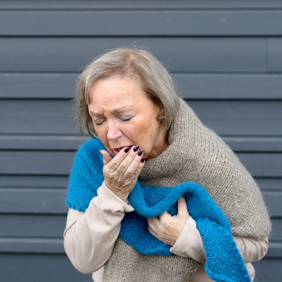 Elegant senior woman coughing into her hand while clutching her chest in a concept of seasonal bronchitis or influenza and medical healthcare grey metal background with copy space
