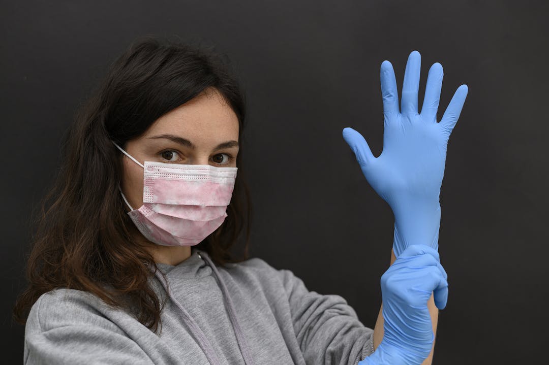 A brunette woman puts on her left hand a blue medical disposable glove. Both hands in gloves. Girl wears a protective mask. A piercing glance at the camera. Remedies for viral infection.
