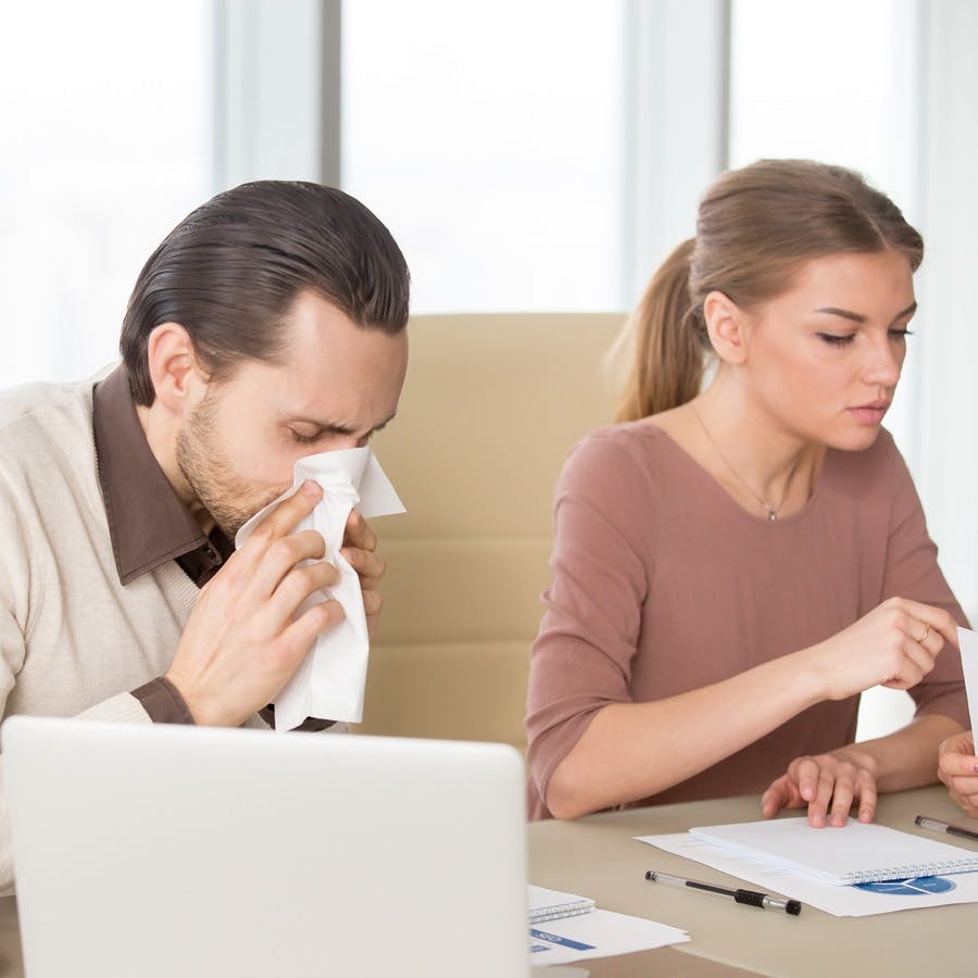 Sick man with handkerchief sneezing blowing nose while working with coworkers, businessman caught cold, seasonal flu. Pandemic influenza, disease prevention, air conditioning in office cause sickness
