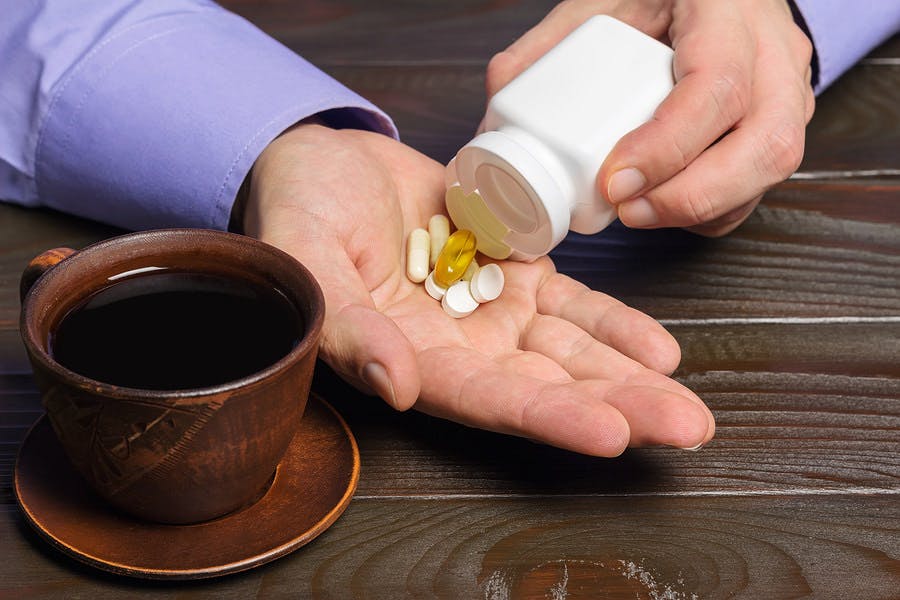 Male hand holds vitamin complex and dietary supplement capsules of omega 3 glucosamine calcium pills cup of black coffee on dark wooden table.
