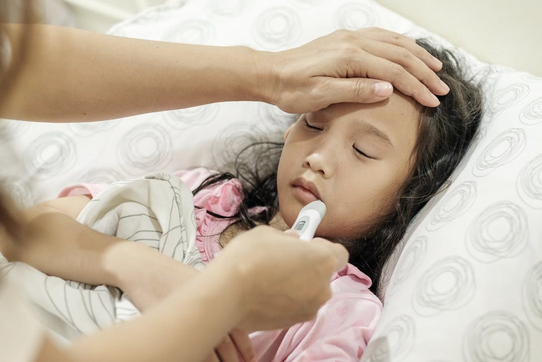 Asian child girl is sick and has a thermometer in her mouth while lying on the bed. Mother checking temperature of her sick daughter with thermometer.  Sick child with fever and illness in bed.
