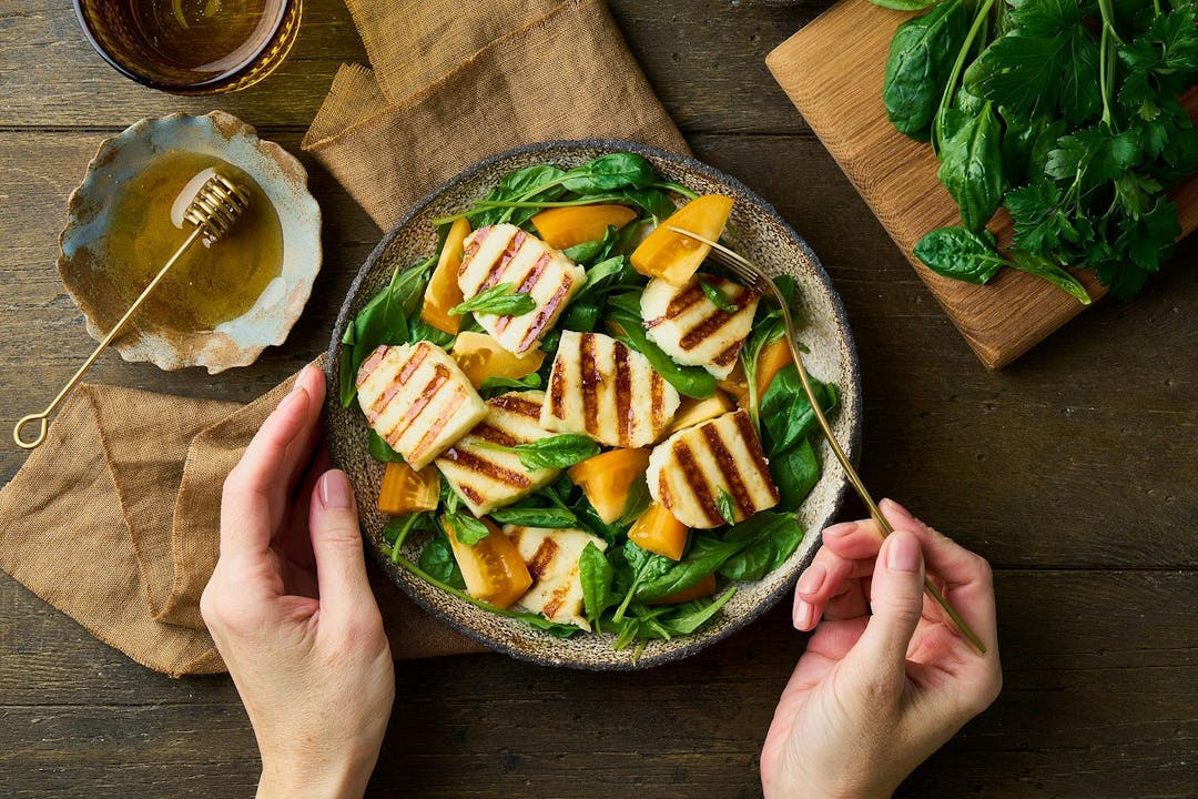 Eating tasty vegetarian healthy diet salad with orange tomatoes, fresh spinach, halloumi cheese and honey. Unrecognisable female hands holding ceramic plate and fork. Wooden background

