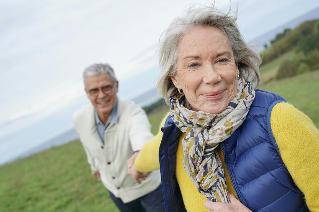Healthy senior woman holding husband&#8217;s hand and leading way on countryside walk

