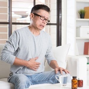 Mature 50s Asian man stomachache,  pressing on stomach with painful expression, sitting on sofa at home, medicines on table.
