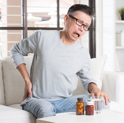 Portrait of casual 50s mature Asian man back pain, pressing on hip with painful expression, sitting on sofa at home, medicines and water on table.
