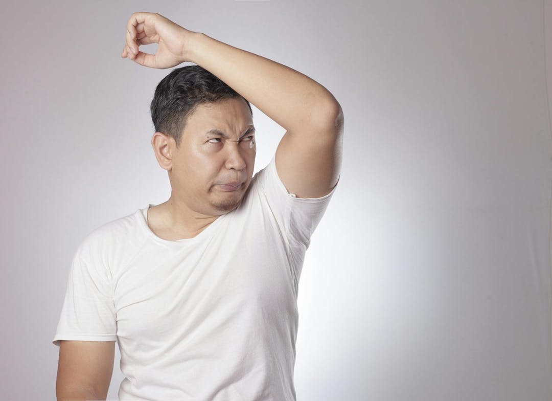 Portrait of funny young Asian man checking his own armpit smell, bad body odor problem
