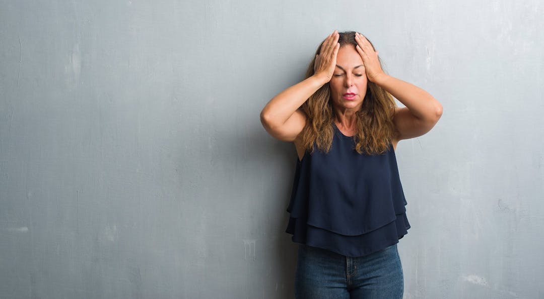 Middle age hispanic woman standing over grey grunge wall suffering from headache desperate and stressed because pain and migraine. Hands on head.
