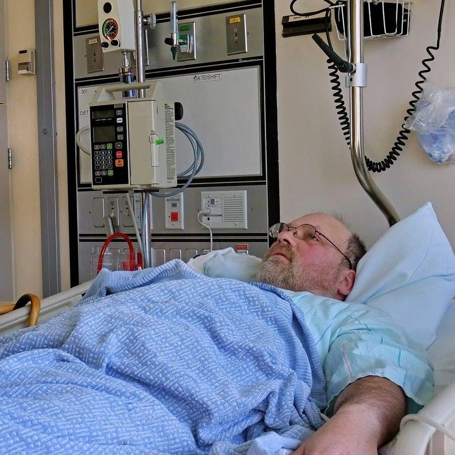 This image is a middle aged bearded Caucasian man who&#8217;s lying in a hospital bed in the hospital with a tray beside the bed and other medical objects.
