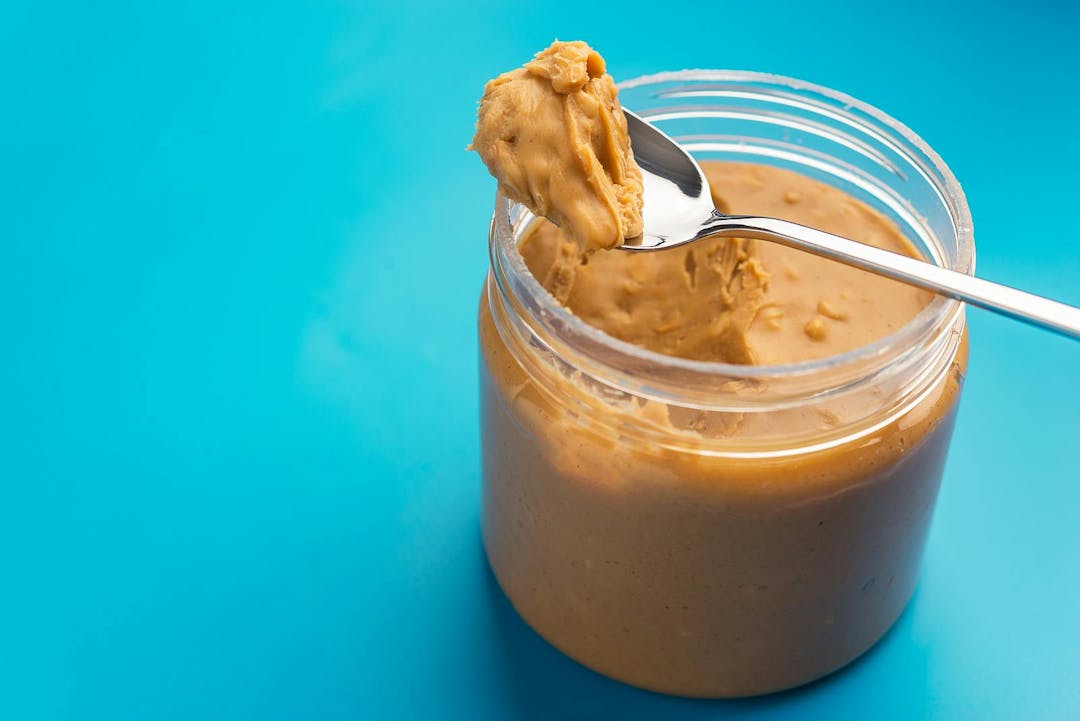Plastic jar with peanut butter and metal spoon on blue background
