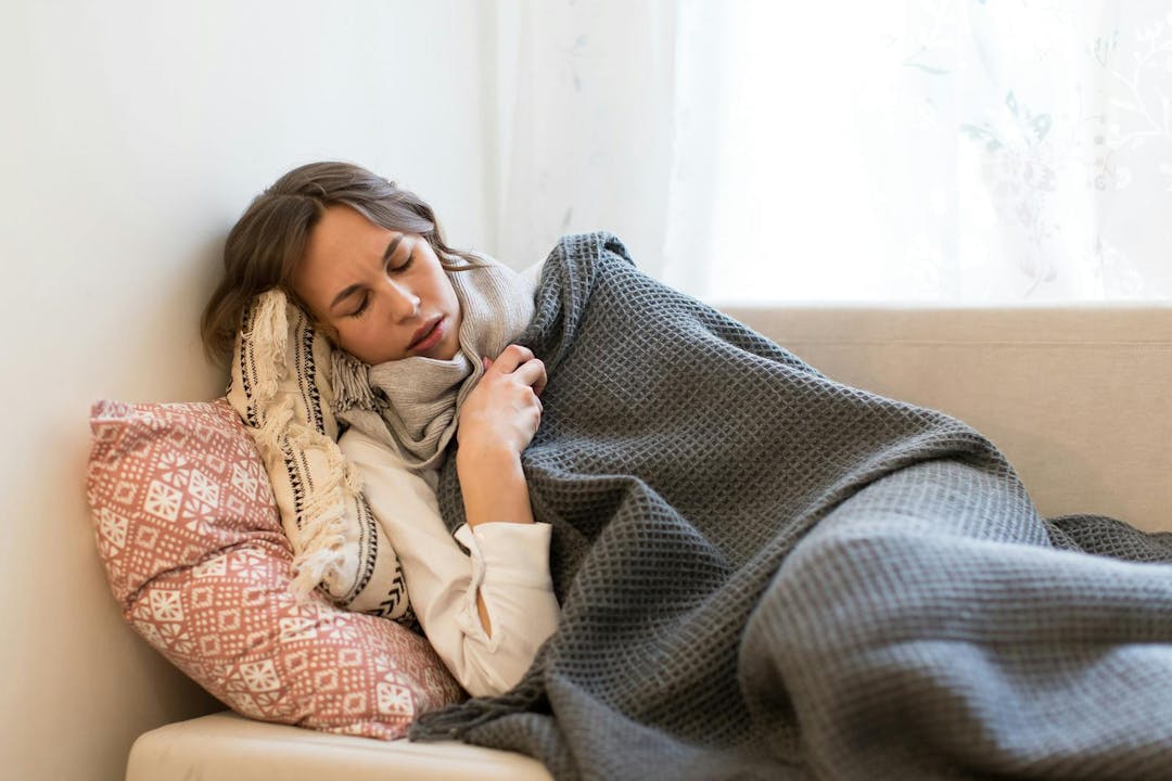 Sick young Caucasian woman lying on sofa and trembles, wrapped in coverlet and scarf, tries to warm herself ,having flu symptoms, feels cold, indoors/Cold, virus, sickness concept/Flu season
