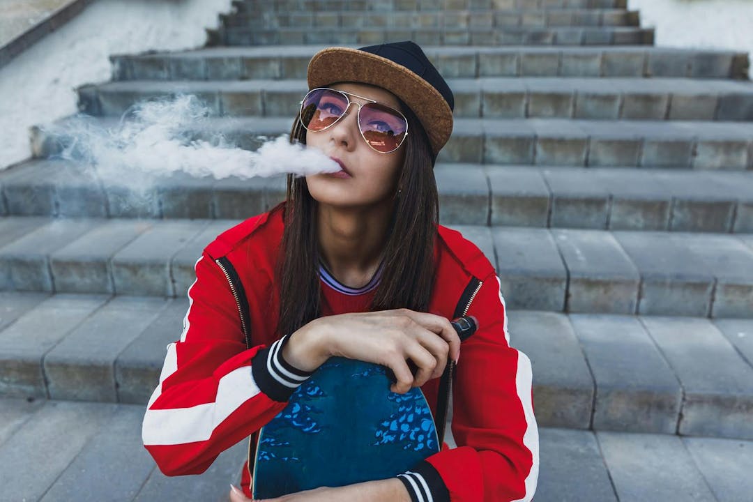 Vaping girl. Young woman with skateboard vape e-cig. Pretty young female in black hat, red clothing vape ecig, vaping device at the sunset. Toned image. Hip-hop style.
