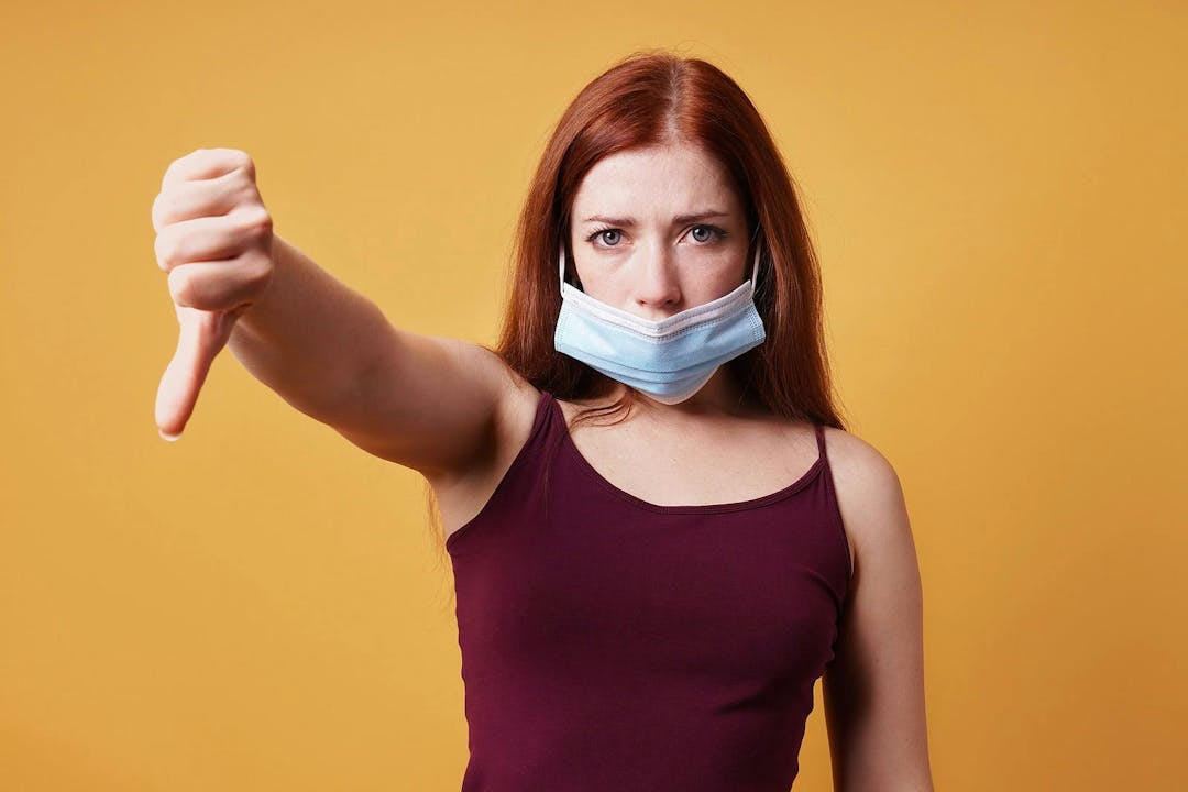 Young woman wearing medical face mask wrong under nose and making thumbs down gesture &#8211; protest against corona virus covid measures
