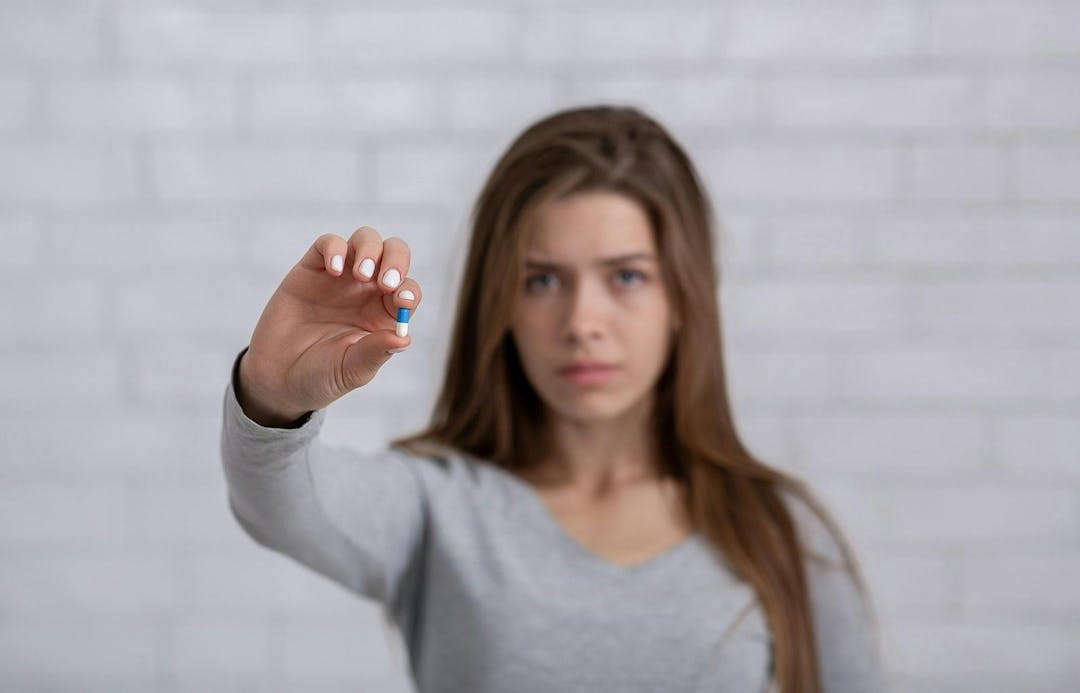 Depressed young lady showing antidepressant or sedative tablet against white brick wall, selective focus. Copy space. Treatment of mental disorders with psychiatric medications
