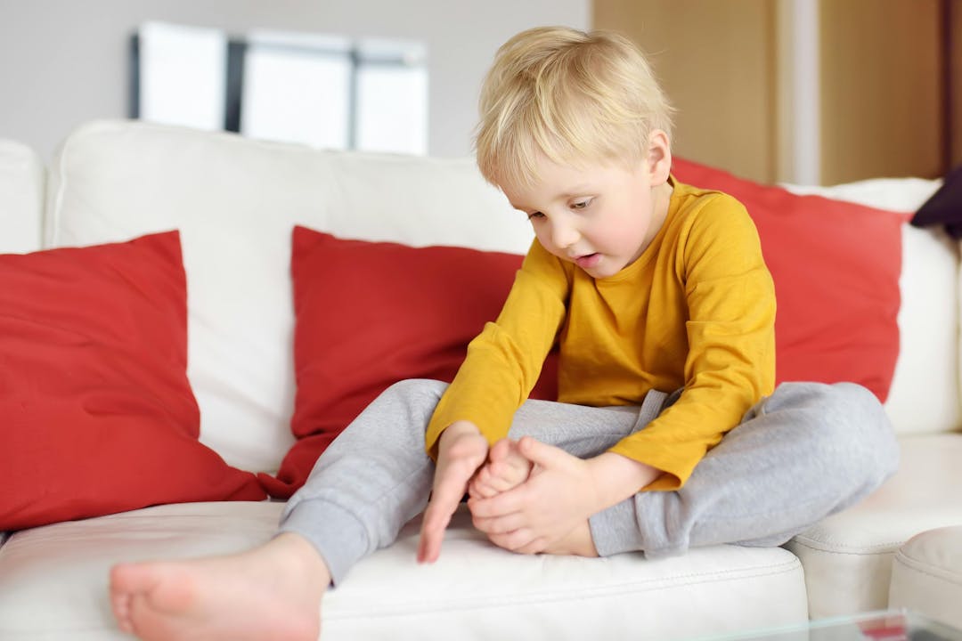 Little boy examines the sore leg sitting on the couch indoors. Injury, bruise, splinter. Kids trauma and safety concepts.
