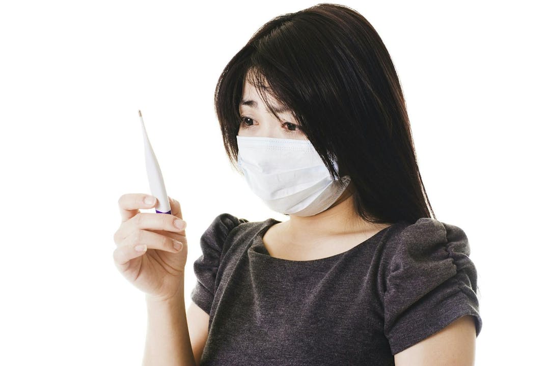 Closeup of a sick Chinese woman wearing a face mask and reading a digital thermometer.
