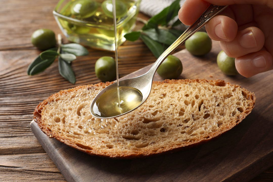 Woman pouring olive oil into spoon over bread at wooden table, closeup
