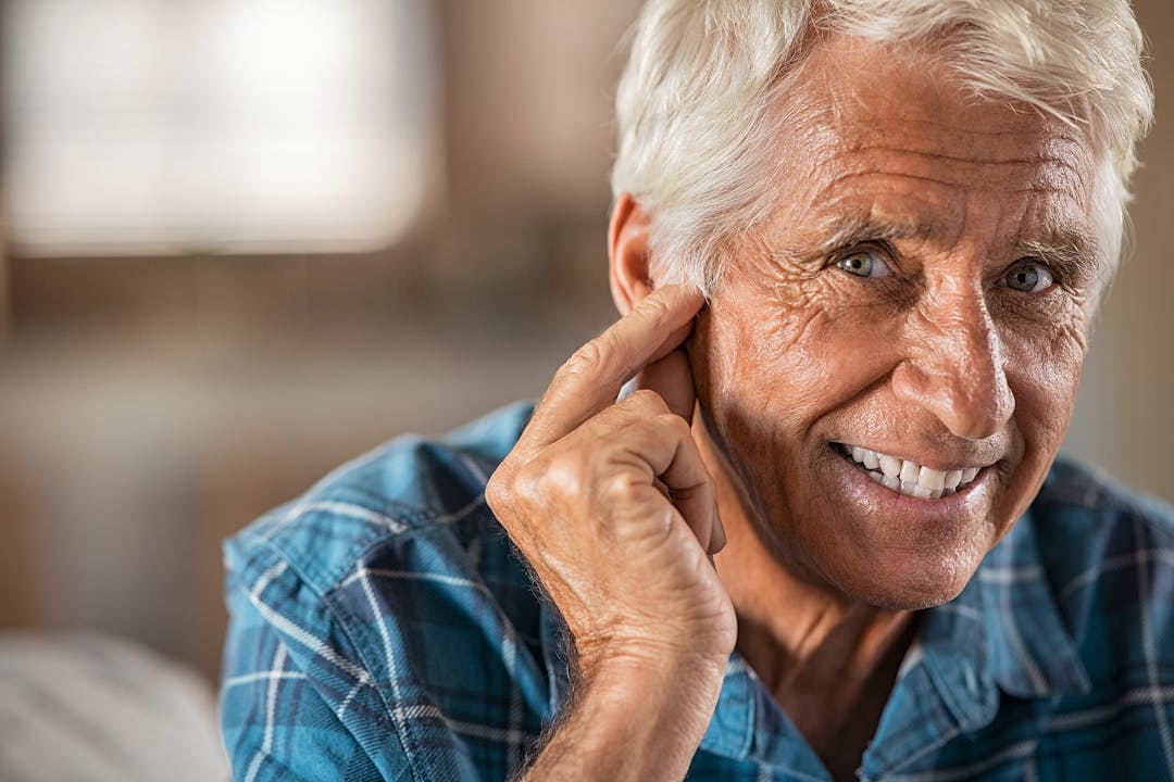 Happy man at home with hearing aid finally hears, copy space. Portrait of smiling senior man holding ear with satisfaction looking at camera. Close up face of old man at home with his new hearing aid.

